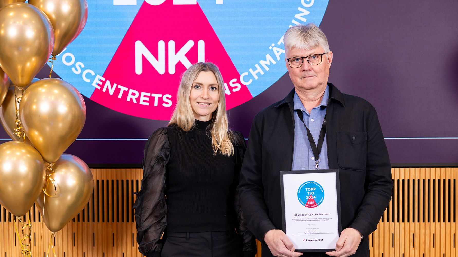 Två personer står framför en bakgrund med en färgglad skylt och ballonger. En av personerna håller ett inramat diplom