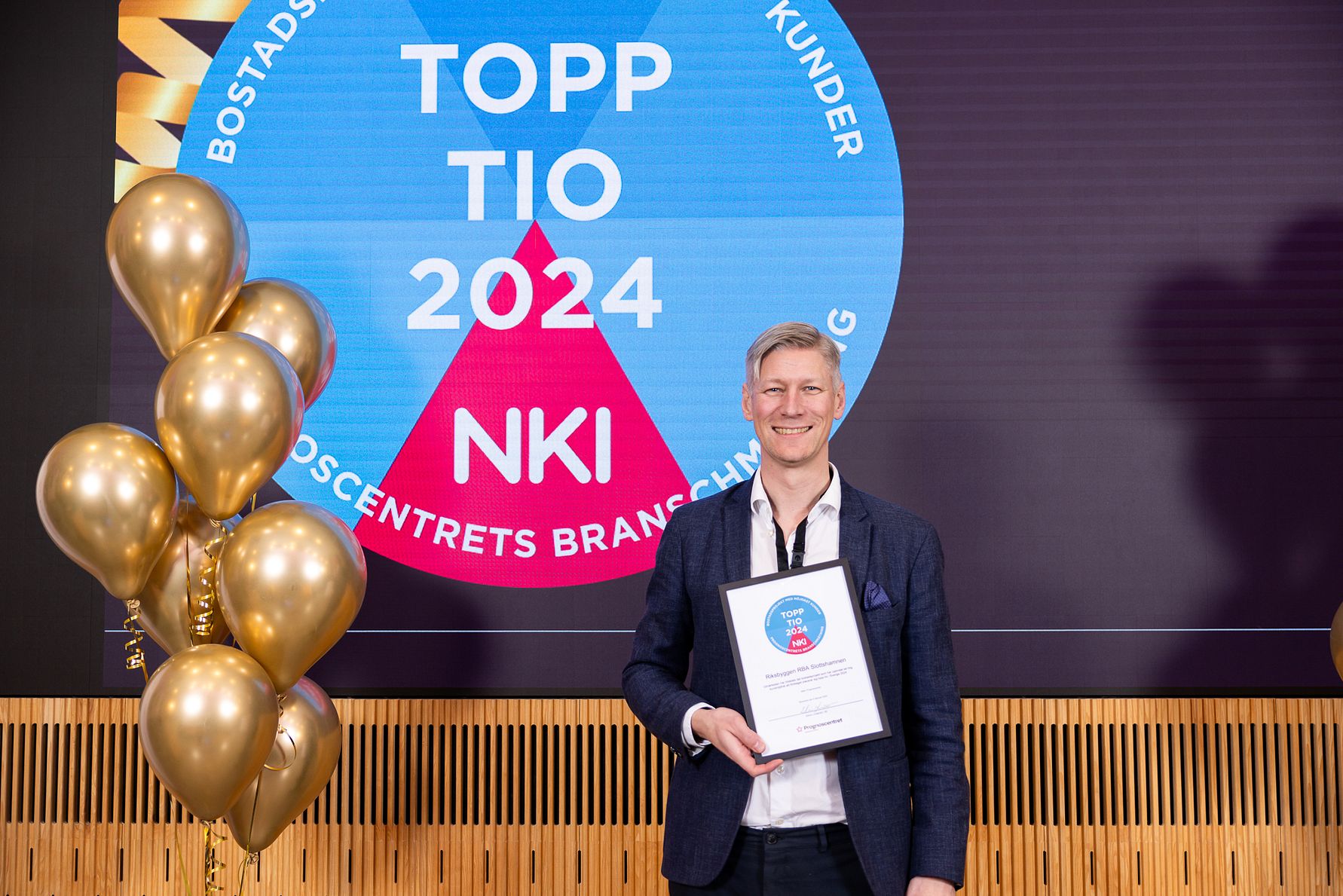 Man i mörk kavaj håller ett diplom framför en skärm med texten "TOPP TIO 2024 NKI" och omgivande guldfärgade ballonger