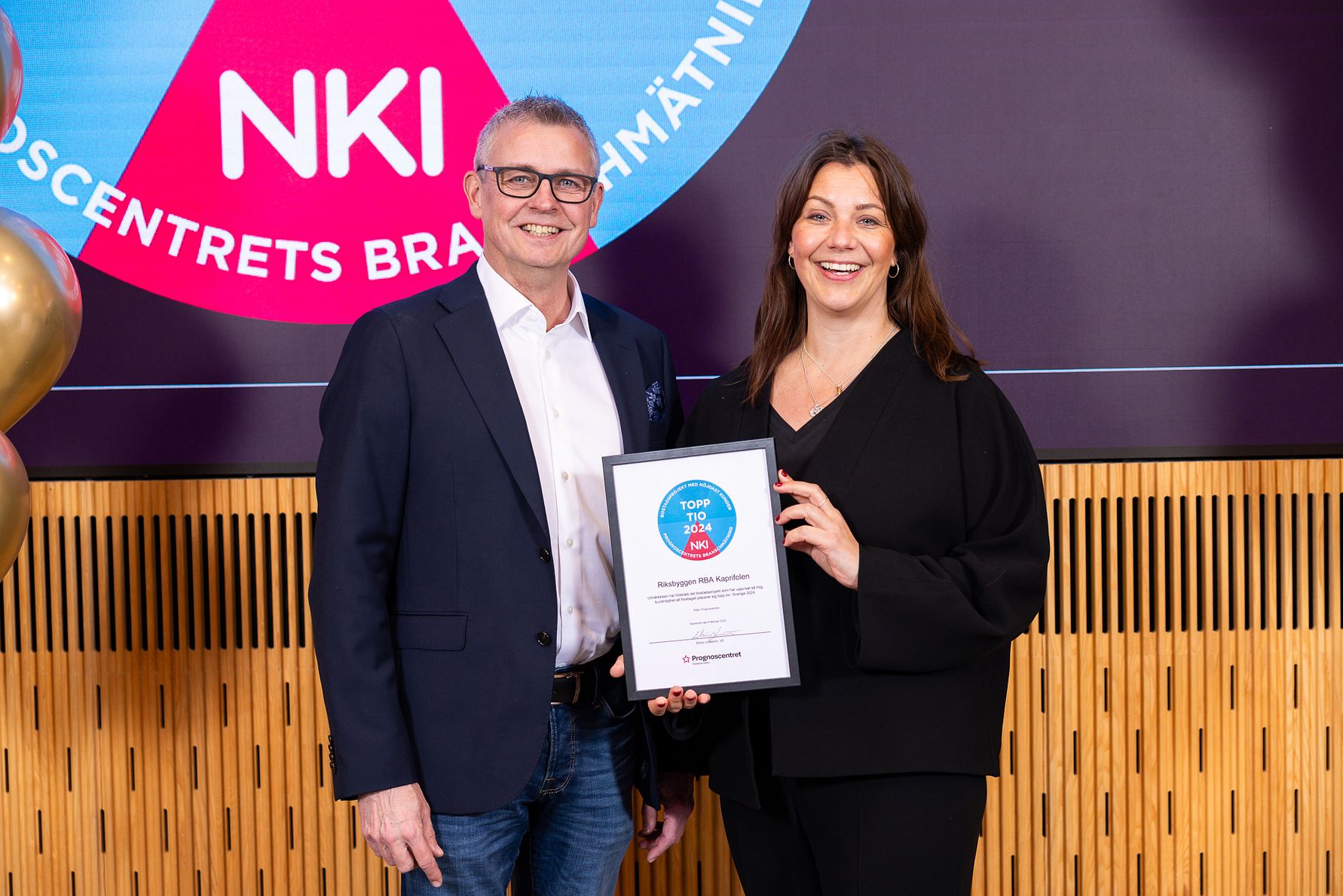 Två personer står framför en bakgrund med en skylt som visar "NKI" och håller ett diplom märkt "Top NKI 2023