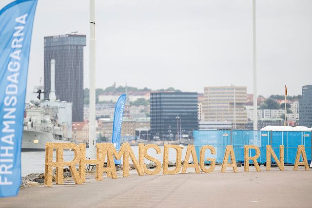 Skylt med texten "Frihamnsdagarna" i halmbokstäver placerad vid en kaj med byggnader och ett krigsfartyg i bakgrunden. Flera blå flaggor syns längs kajen