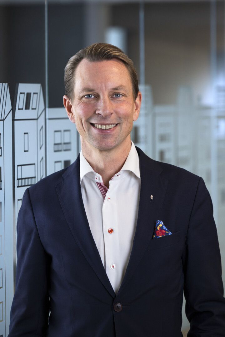 En man i mörk kostym och vit skjorta med röda knappar står framför en glasvägg med ett mönster av byggnader