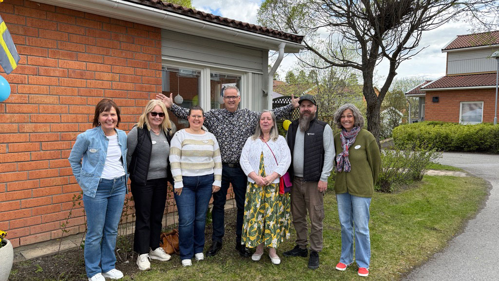 Grupp av människor utanför ett tegelhus, med träd och gräsmatta i bakgrunden