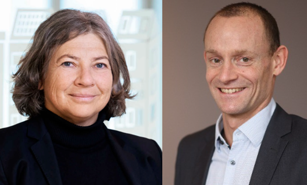 Två personer iförda kostymer står inomhus; den ena har mörkt långt hår och den andra kortklippt hår, mot suddiga bakgrunder