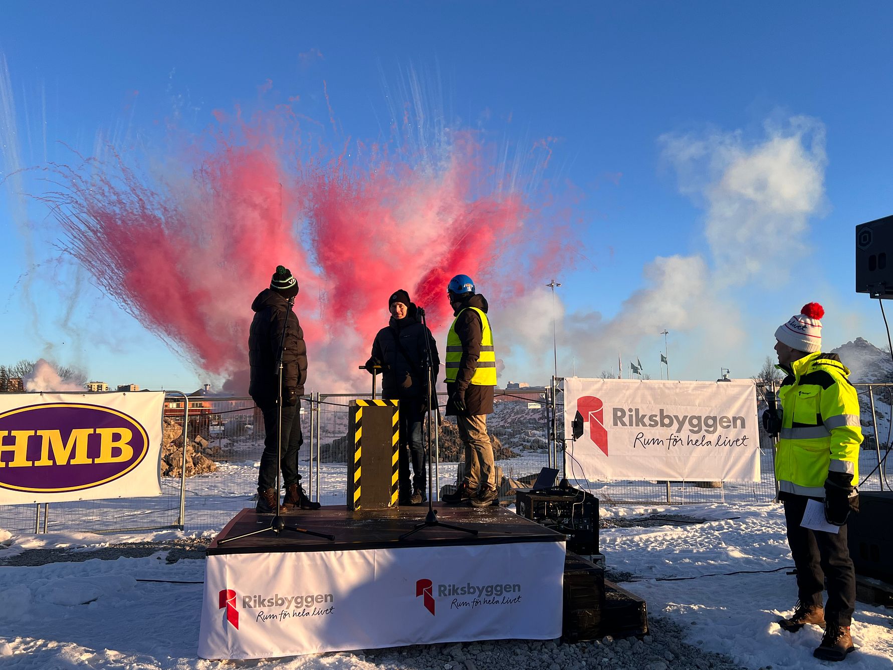 Fyra personer står på en liten scen utomhus omgivna av snö. Bakgrunden visar röda fyrverkerier och banderoller med företagslogotyper