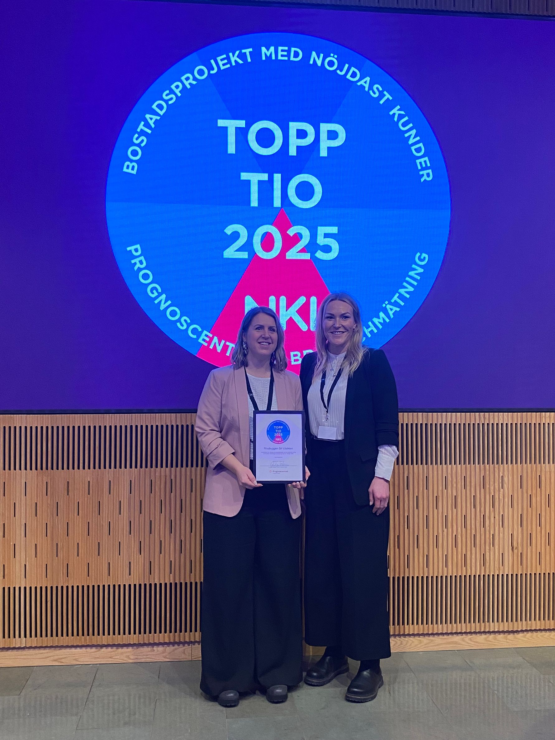 Två personer står framför en skylt med texten "Topp Tio 2025 NKI" och håller i ett diplom. Bakgrunden består av en lila vägg och ett trägolv