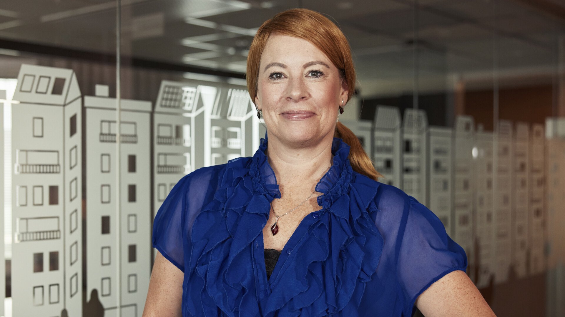 En kvinna i blå blus står framför en glasvägg med kontorsinredning i bakgrunden