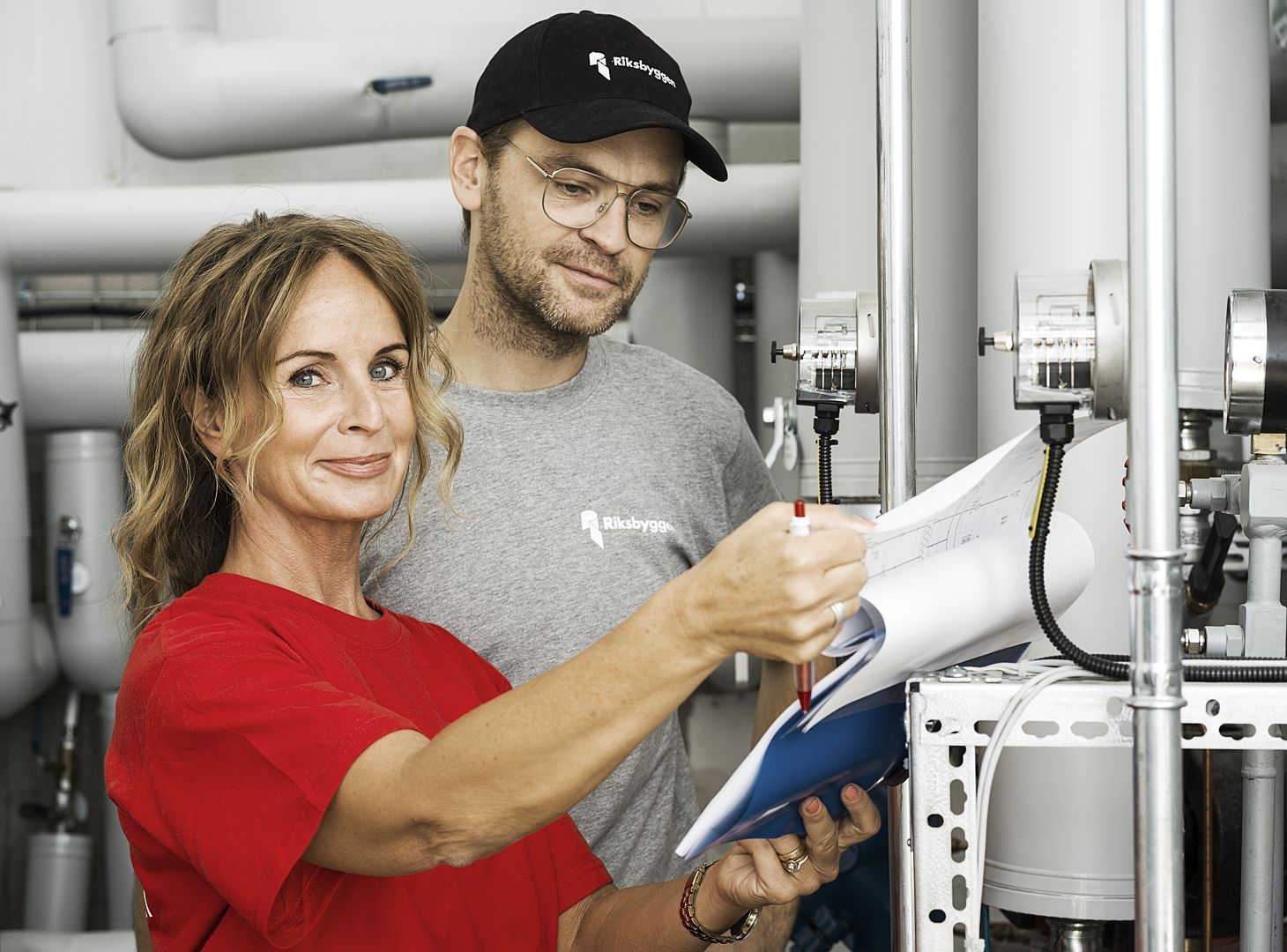 Två personer granskar ritningar vid industriella rör och mätinstrument, en av dem pekar med en penna