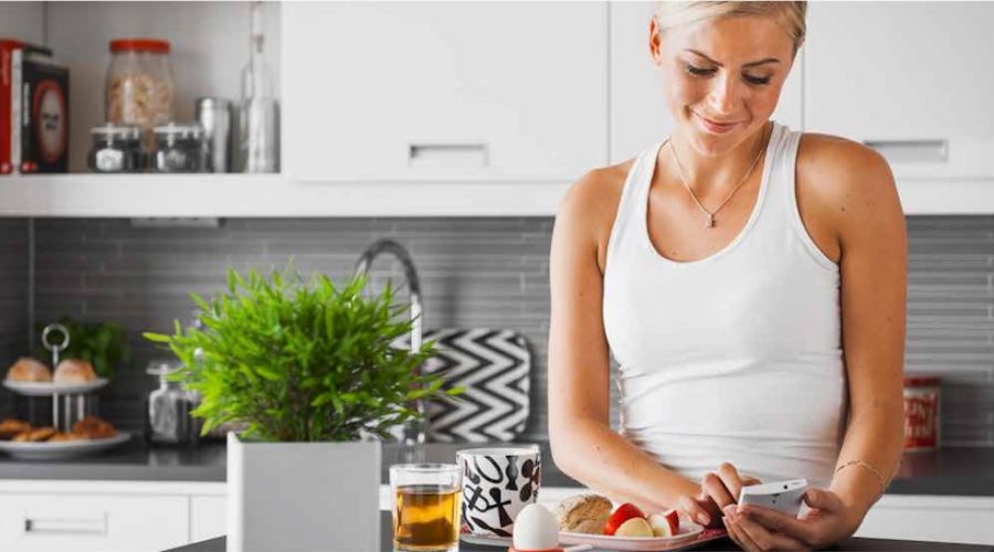 En person i ett modernt kök lutar sig mot en köksbänk med en frukosttallrik, ett kokt ägg, frukt, te och en smartphone. På bänken står en grön växt och köksredskap i bakgrunden