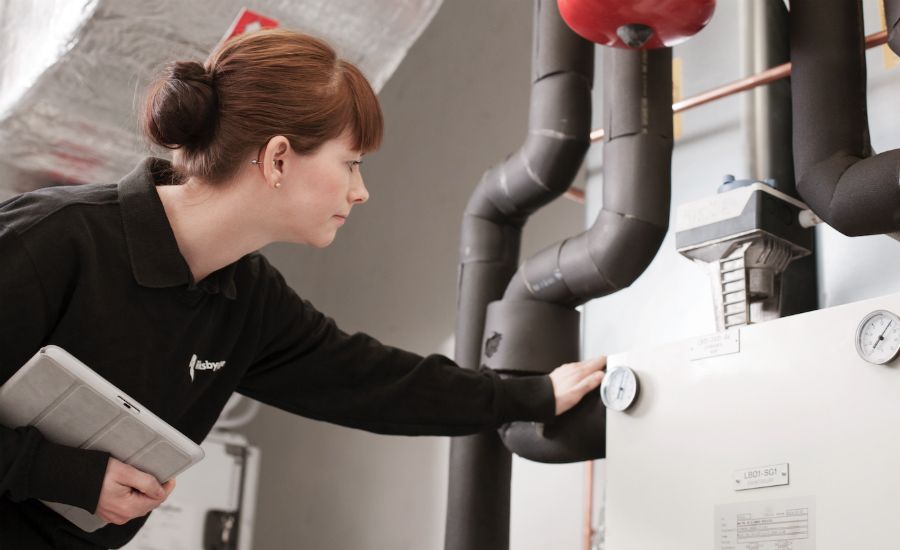 Kvinna i arbetskläder inspekterar en vit industriell maskin med mätare, omgiven av isolerade rör och ventilationssystem