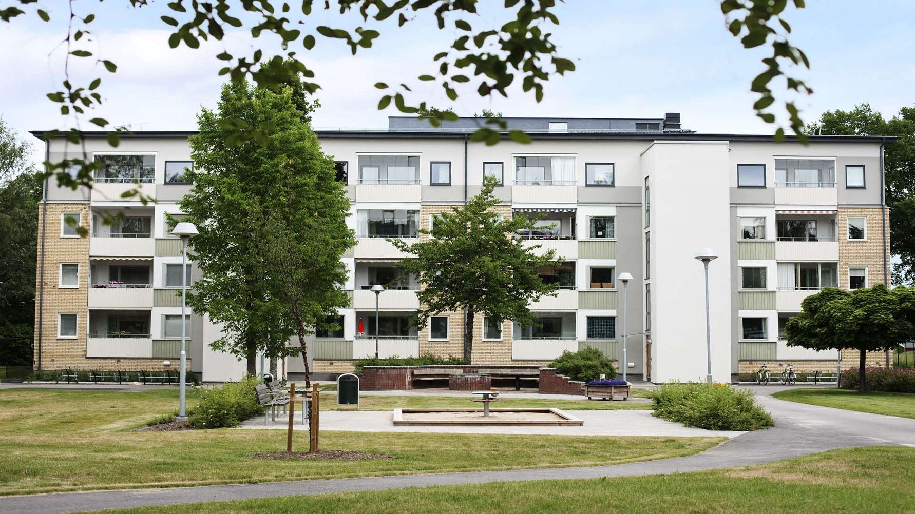 Flerbostadshus i modern stil med grönområde, träd och en lekplats i förgrunden under en klar himmel