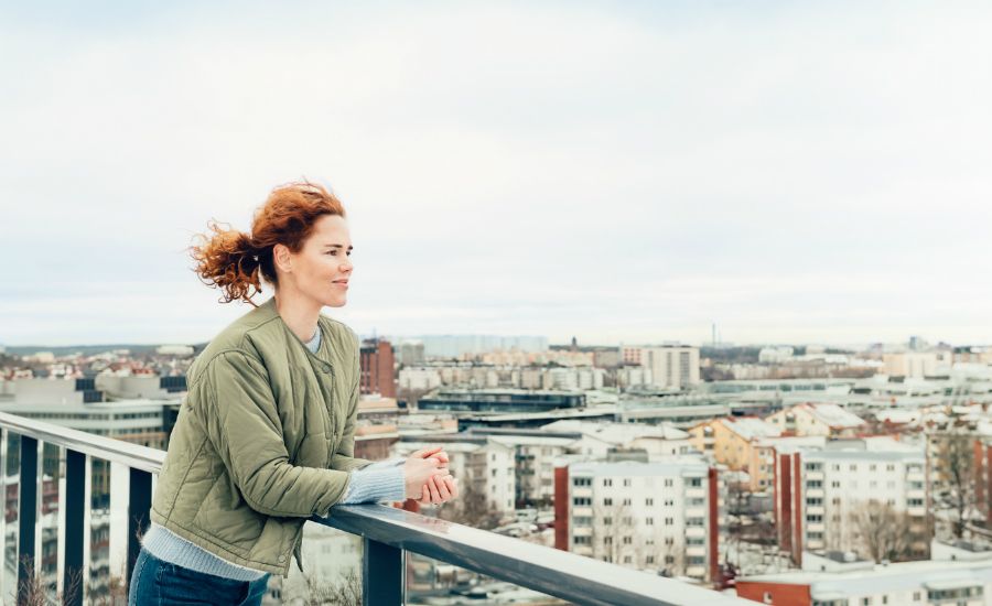 Person med lockigt rött hår lutar sig mot ett räcke och blickar ut över en stad med byggnader och molnig himmel i bakgrunden