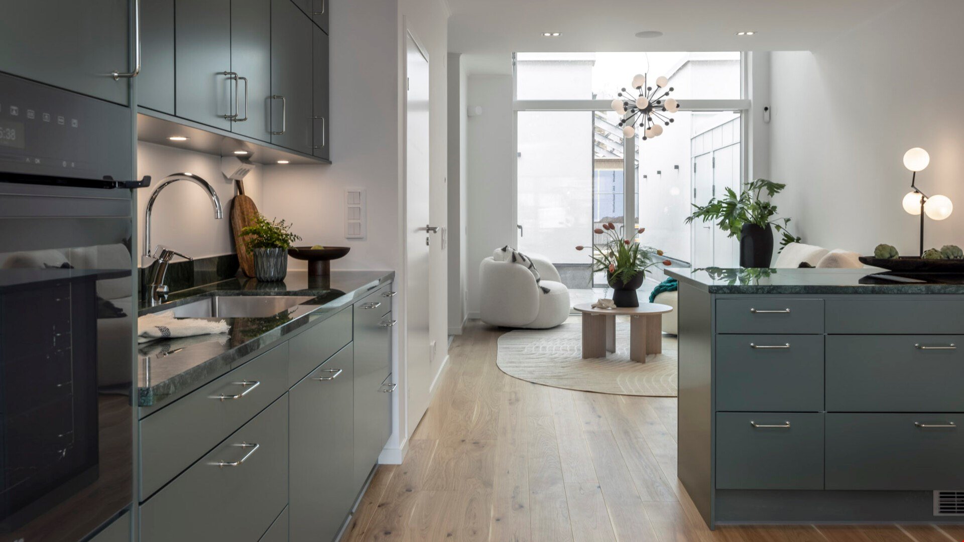 Modernt kök och vardagsrum med gråa skåp, rostfri diskho, trägolv och stora fönster som släpper in naturligt ljus