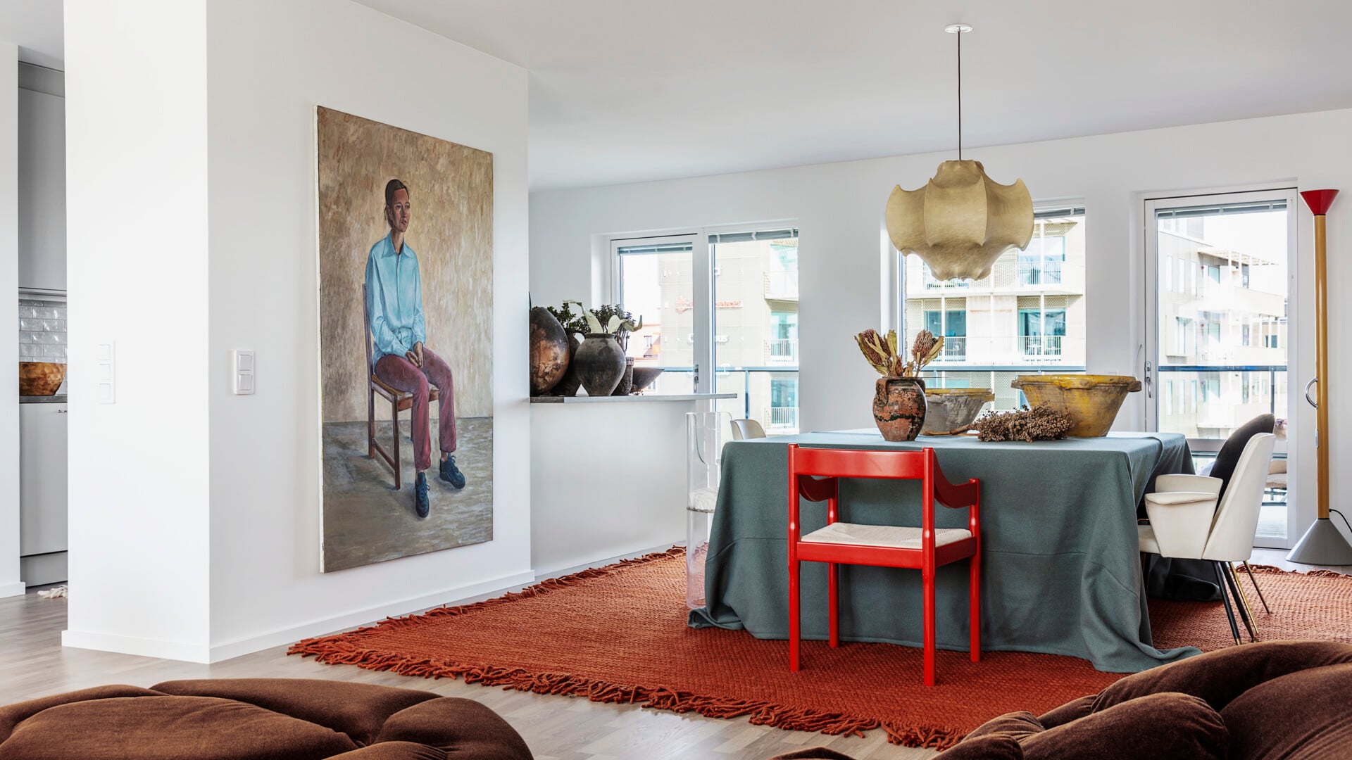Modernt vardagsrum med en stor målning av en person på en stol, ett matbord med en röd stol och olika dekorativa föremål, samt stora fönster i bakgrunden