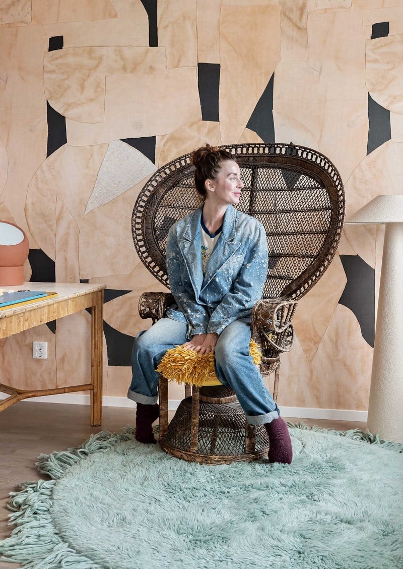 Marie Olsson Nylander i jeansjacka och jeans sitter på en rottingstol i ett rum med ljusblå matta och mönstrad vägg