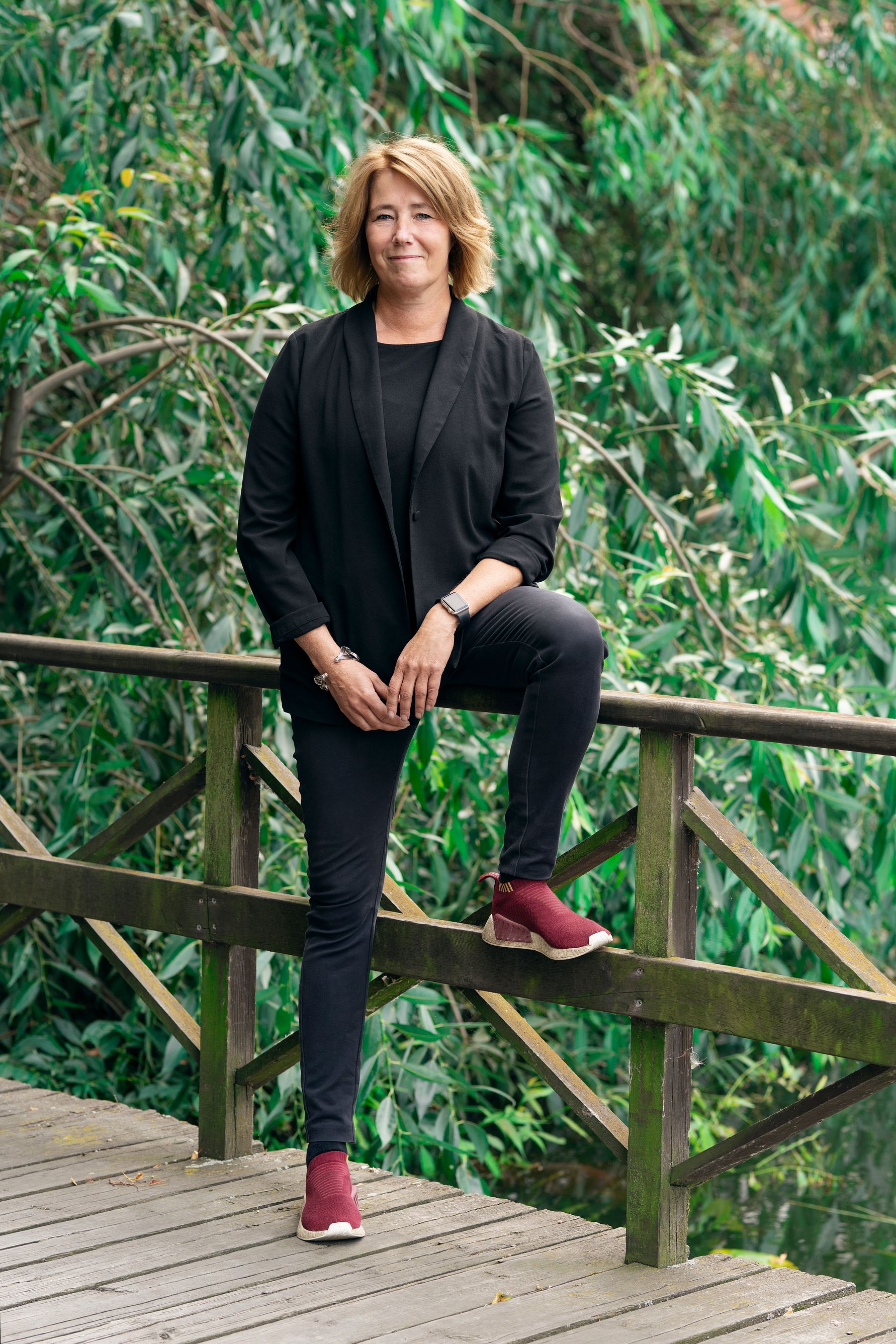 Portrait of a woman in black clothing standing on a wooden bridge in a green outdoor setting