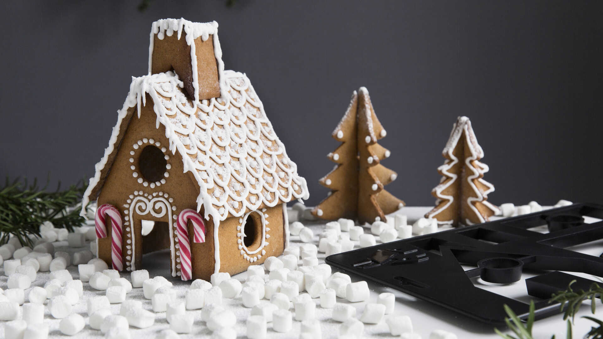 Pepparkakshus dekorerat med kristyr och polkagrisar, omgivet av små marshmallows och pepparkaksgranar på ett bord