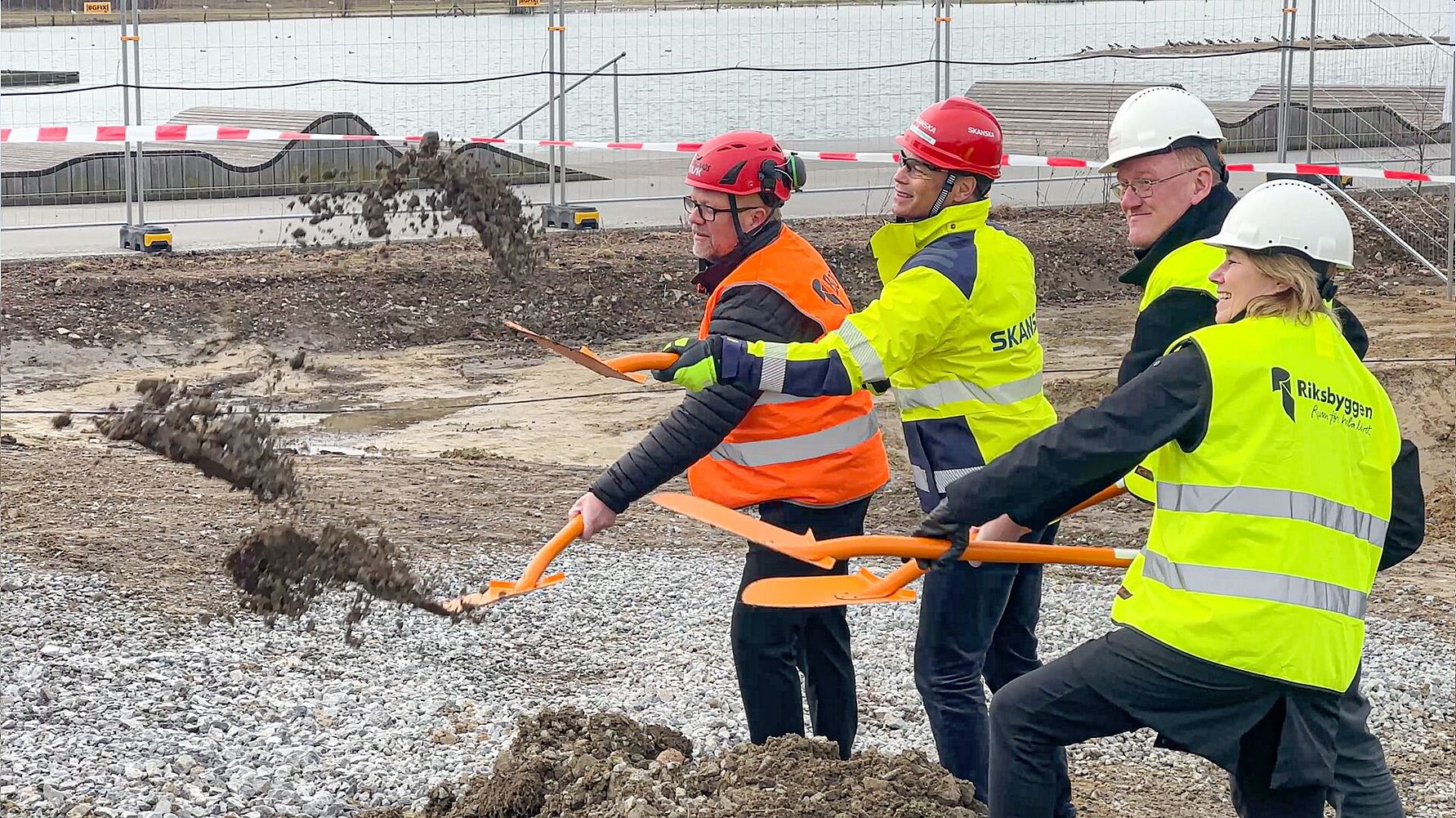 Fyra personer i hjälm och varselkläder spadar jord på en byggarbetsplats