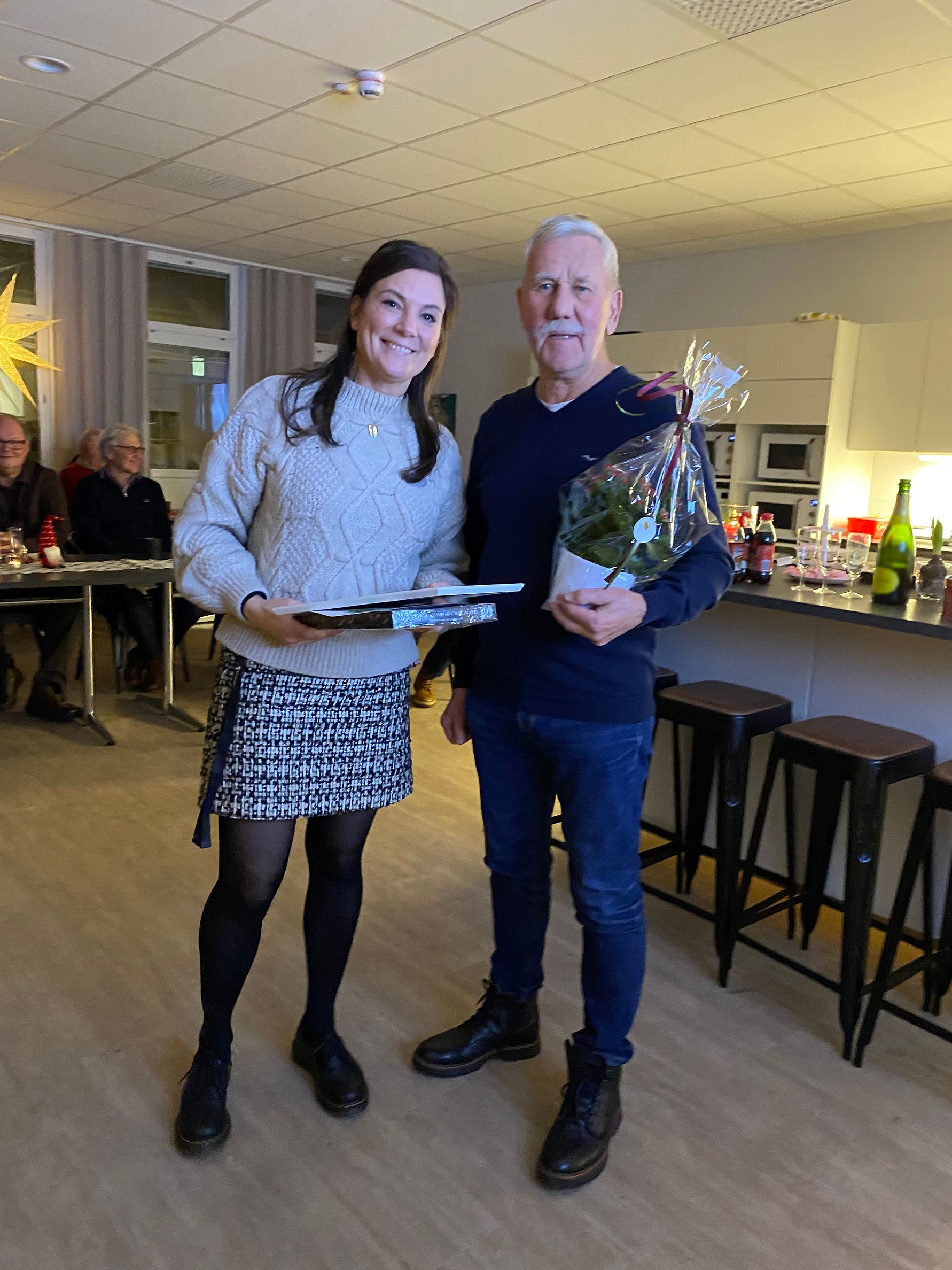 Två personer står inomhus i ett upplyst rum, där den ena håller en blombukett inslagen i cellofan och den andra håller en bok eller mapp. Bakom dem syns stolar, bord och en köksmiljö