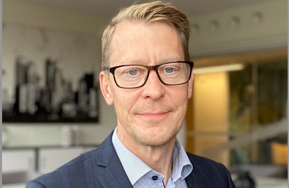 En man i kavaj och skjorta står i en kontorsmiljö med suddig bakgrund som visar ett stadsmotiv och ljusa väggar