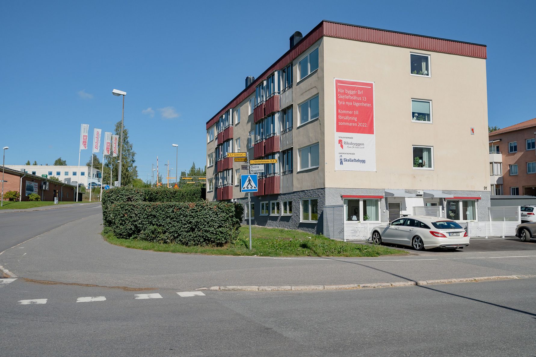 Flerfamiljshus med beige och röda fasader, en skylt med text på väggen, parkerade bilar och en trottoar vid en korsning
