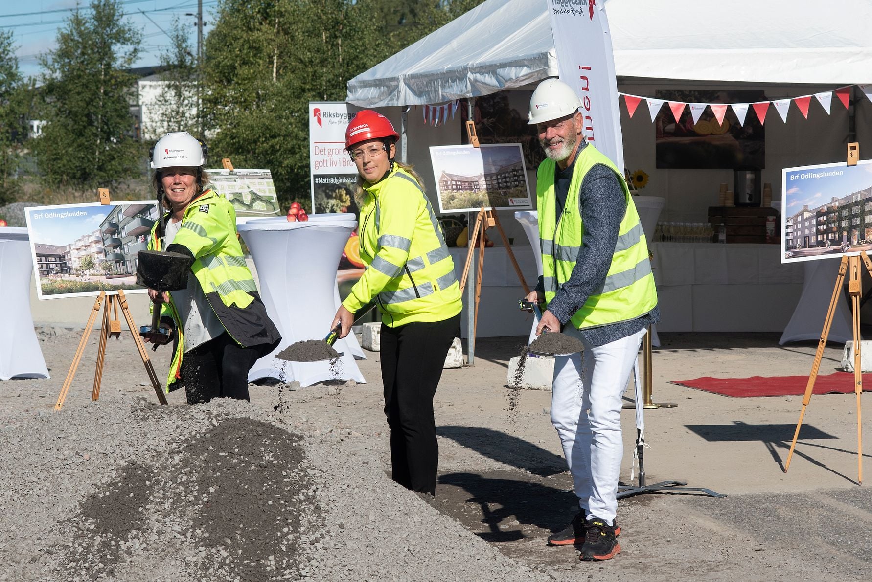Tre personer i reflexvästar och hjälmar skyfflar sand vid en ceremoni på en utomhusplats, med tält och informationsskyltar i bakgrunden