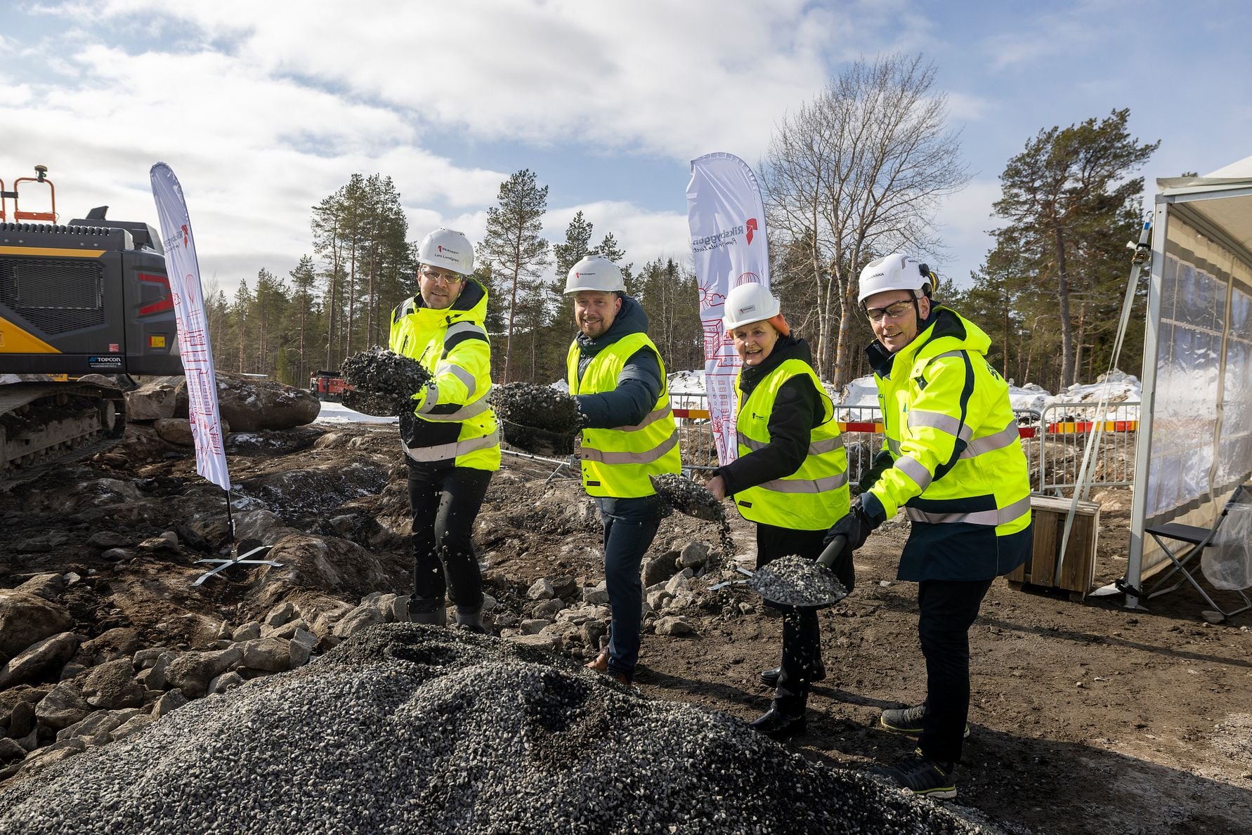 Personer i skyddshjälmar och reflexjackor arbetar med grusläggning på en byggarbetsplats utomhus, omgiven av maskiner, träd och en tältstruktur