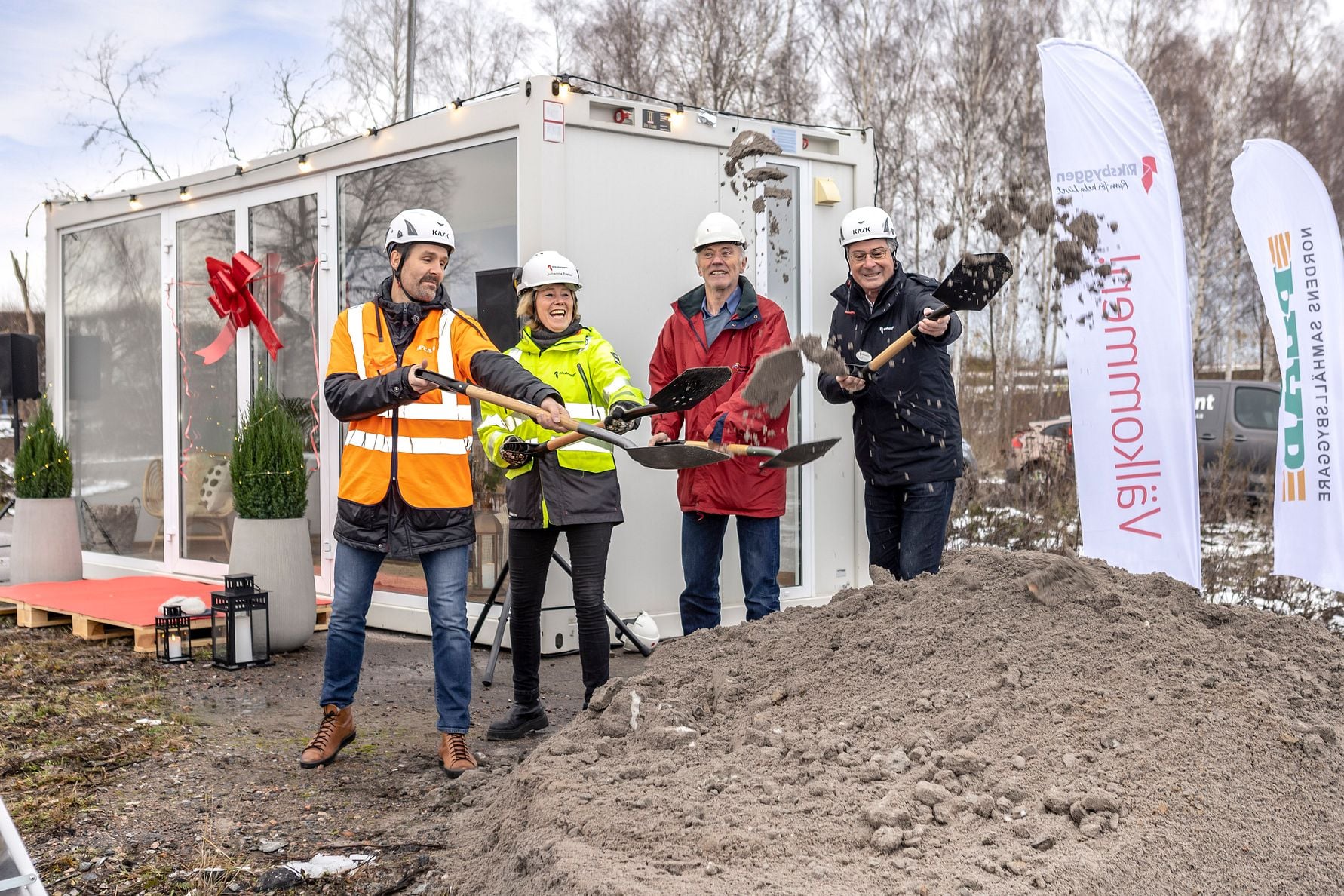 Fyra personer i skyddshjälmar och reflexkläder håller i spadar vid en symbolisk första spadtagsceremoni framför en byggmodul och skyltar