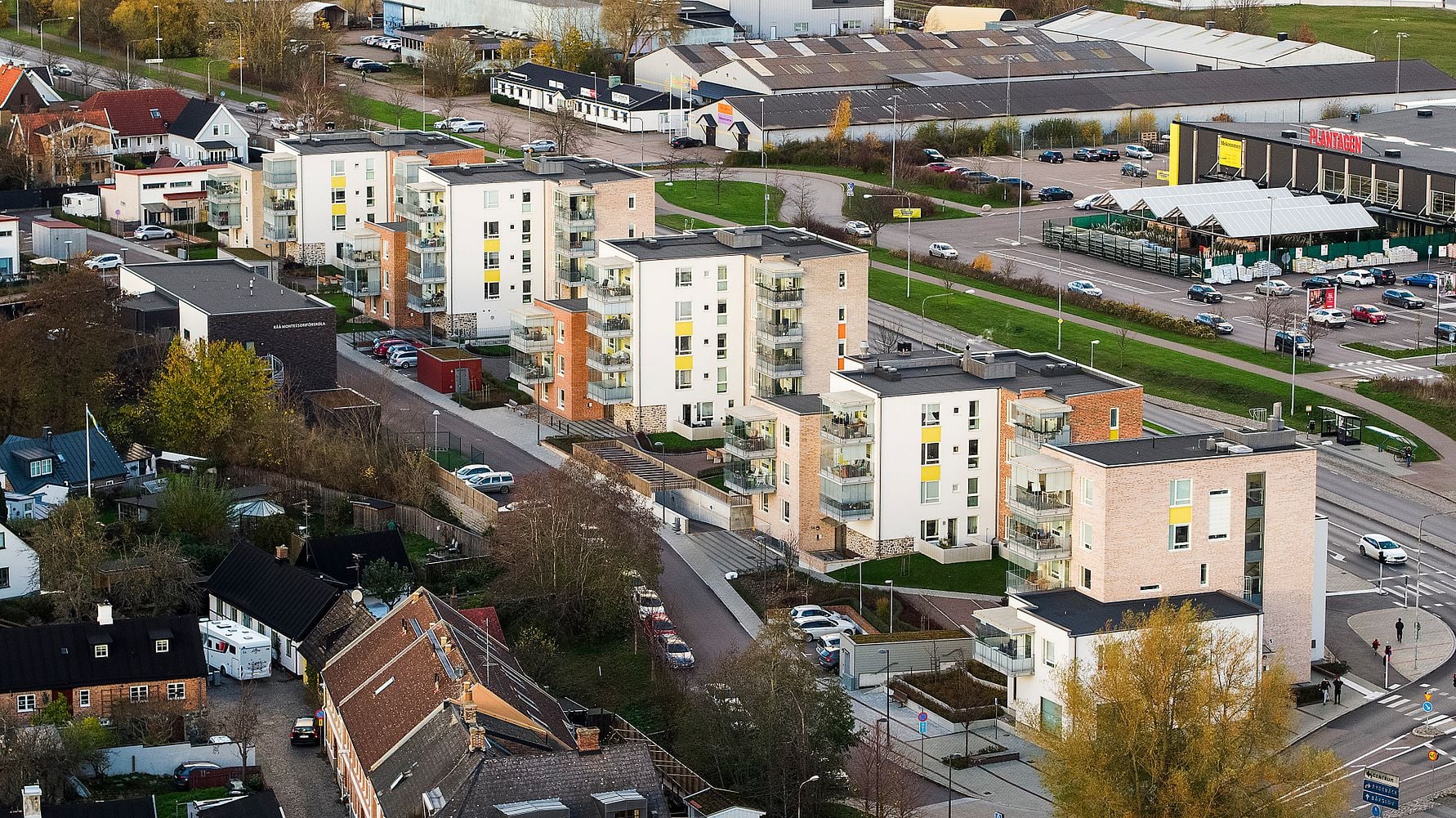 Flygfoto av ett bostadsområde med flerfamiljshus och parkeringsplatser, omgiven av mindre byggnader och en handelsplats i bakgrunden
