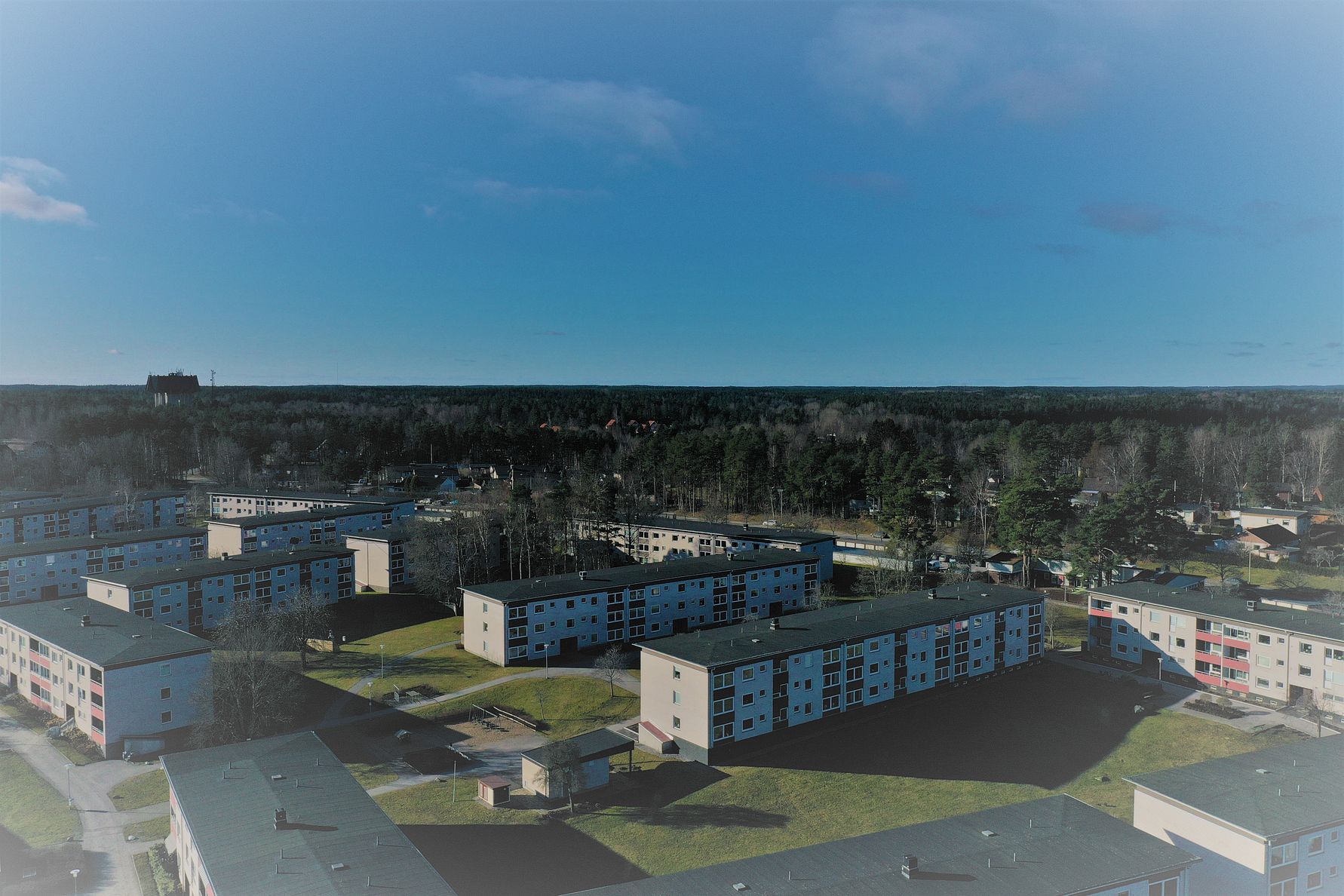 Luftbild av ett bostadsområde med flerfamiljshus, grönområden och skog i bakgrunden under en klarblå himmel