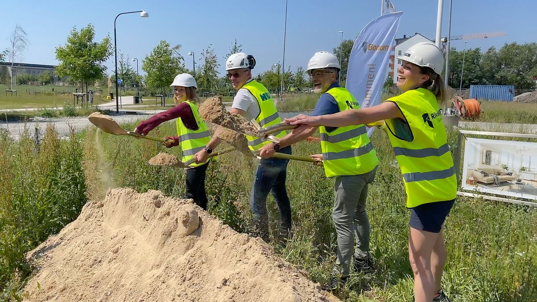 Flera personer iklädda reflexvästar och byggarbetsplats-hjälmar använder spadar för att gräva i en hög med sand på en gräsyta, med byggutrustning och flaggor i bakgrunden