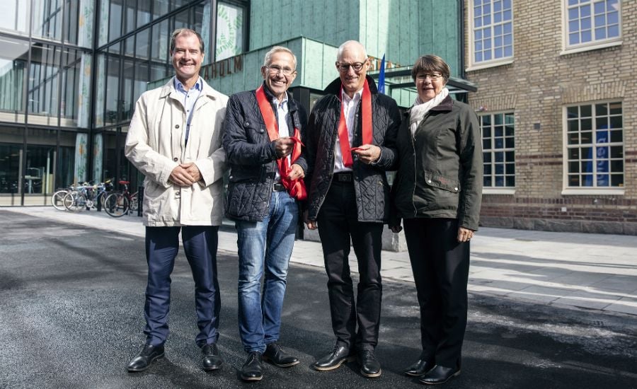 Fyra personer står utomhus framför en modern byggnad och en tegelvägg, med cyklar synliga i bakgrunden. Två av dem bär röda halsdukar