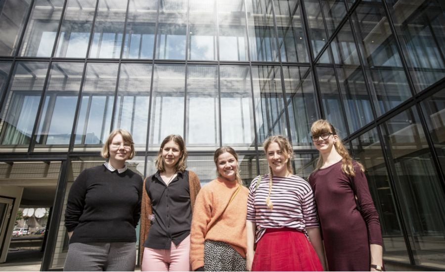 Fem personer står framför en modern glasbyggnad under en klar himmel