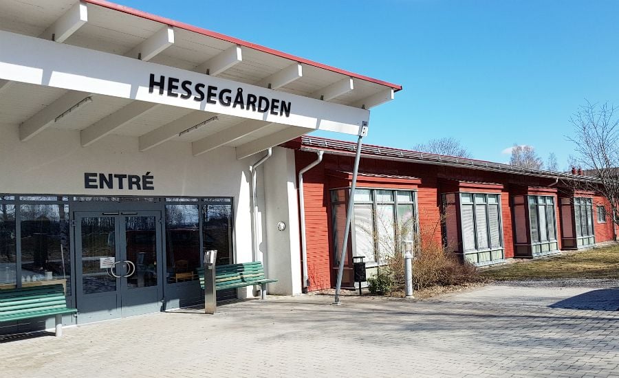 Entré till byggnaden Hessegården med glasdörrar, vita väggar och röda detaljer, omgiven av utomhusområde med bänkar och blå himmel