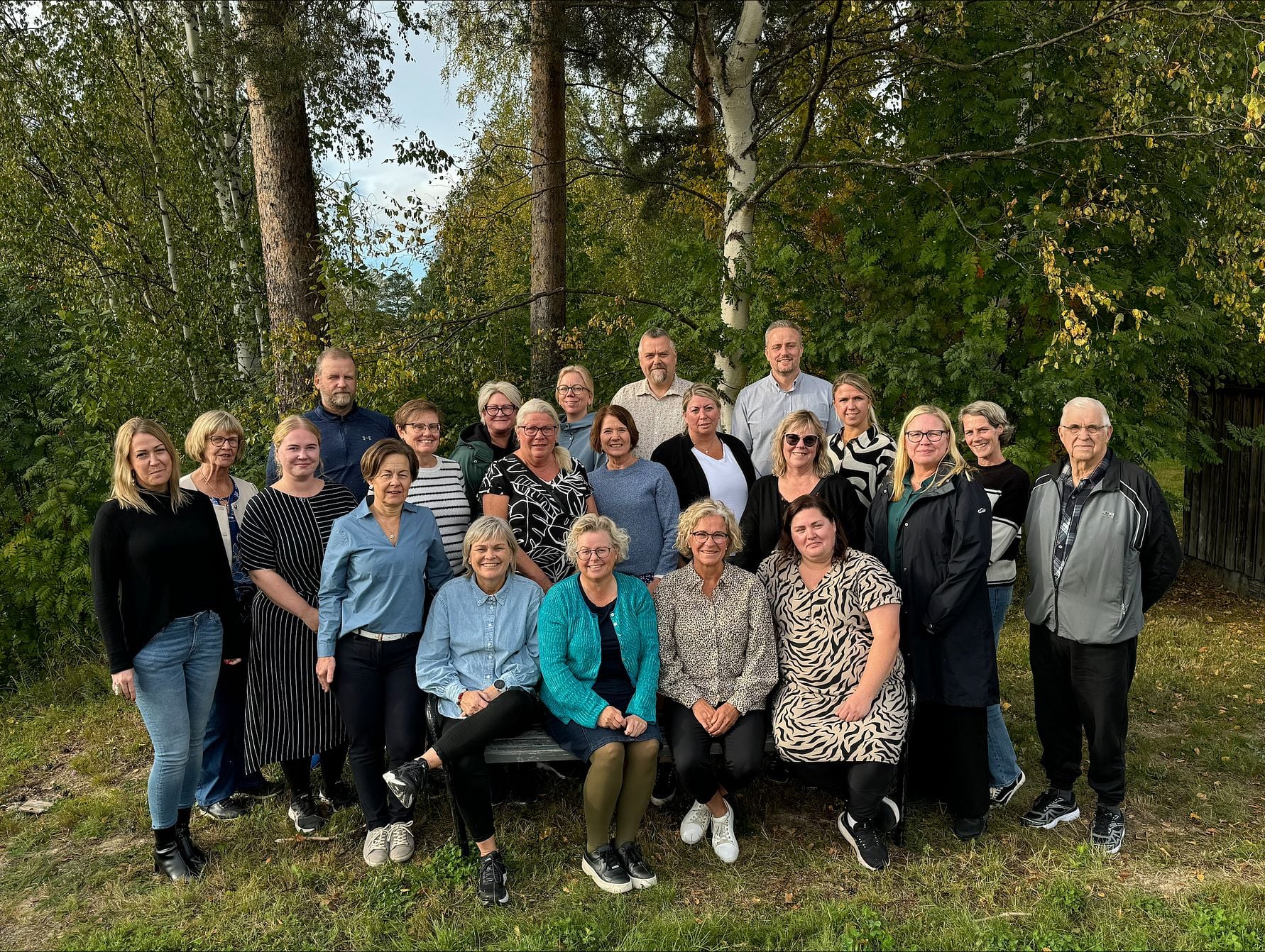 En grupp människor poserar utomhus framför träd och grönska, några sitter på en bänk medan andra står bakom