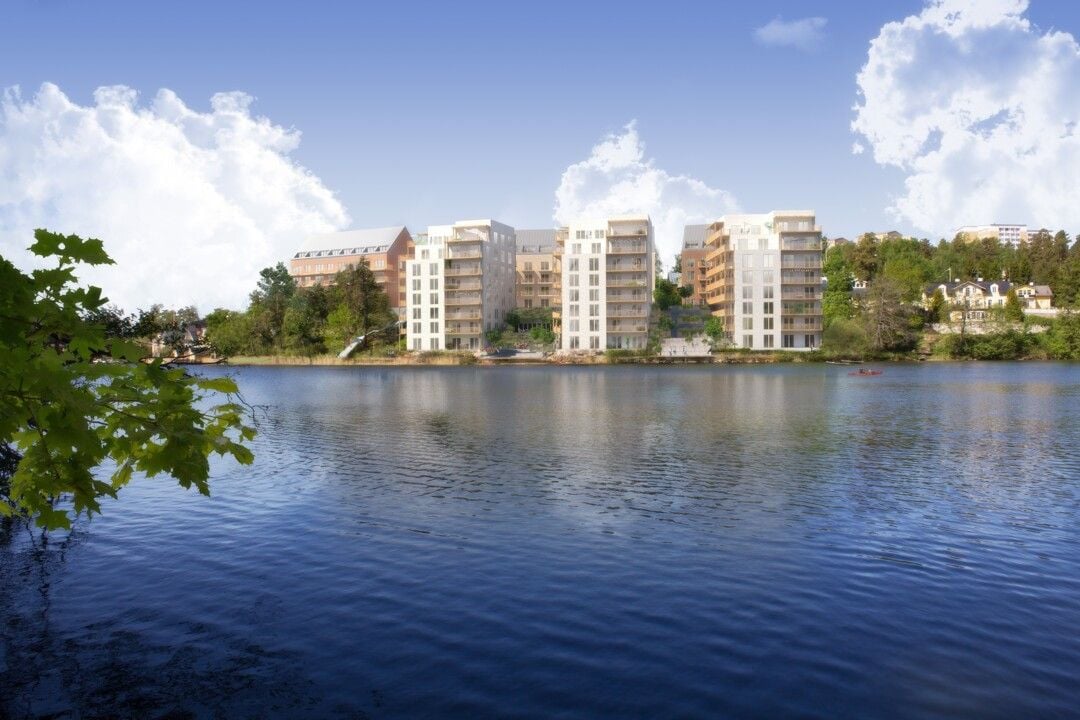 Vy över en sjö med moderna bostadshus och grönområden i bakgrunden under en klar blå himmel