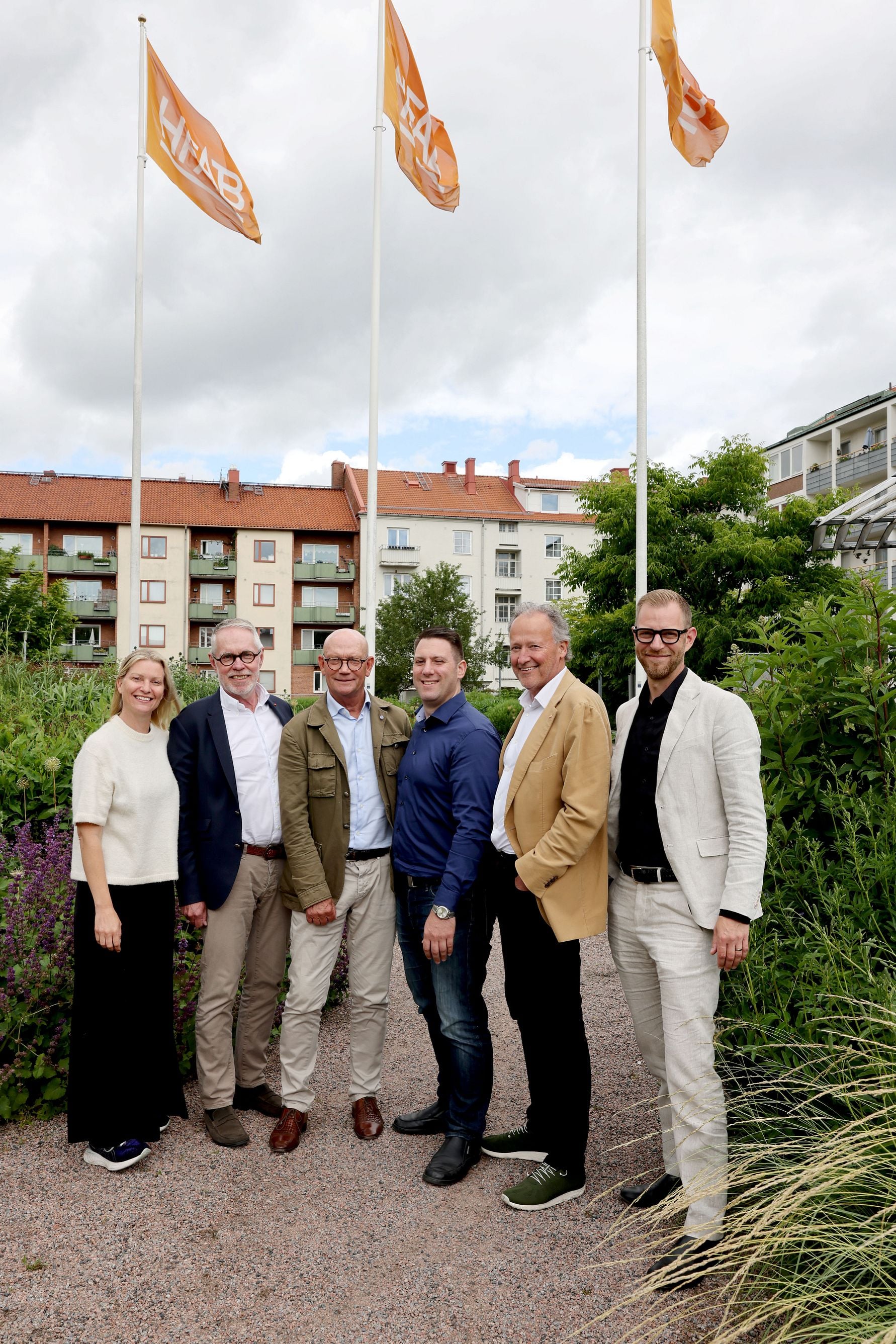 En grupp människor står ute i en trädgård med flaggor i bakgrunden, omgivna av grönska och byggnader