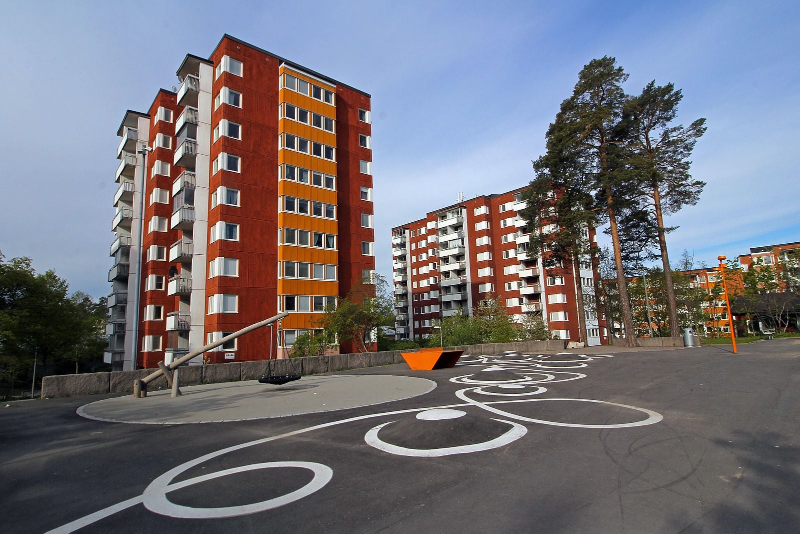 Flerbostadshus med röda fasader och vita balkonger, omgivna av träd och en lekplats med asfaltmönster