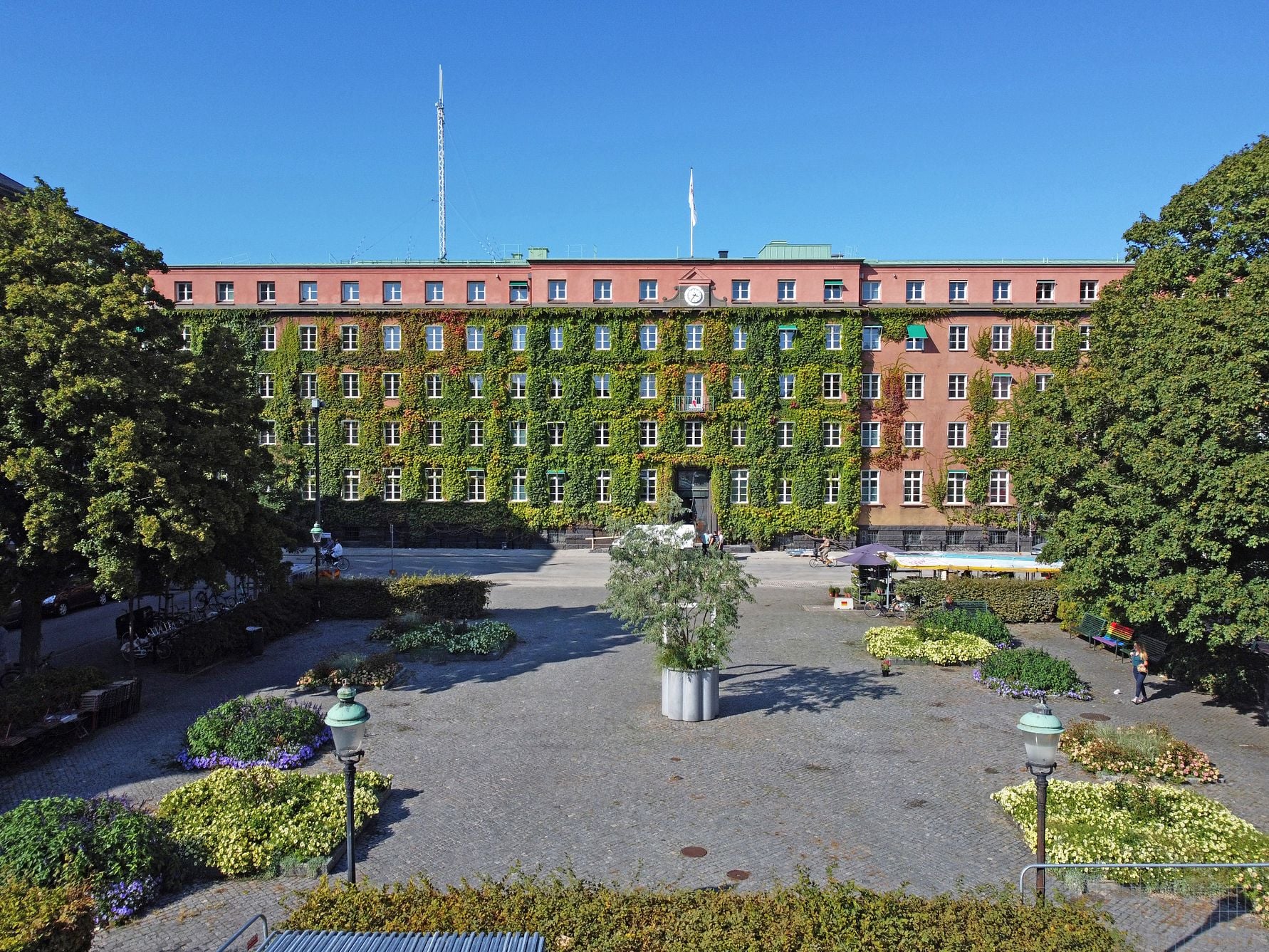 Byggnad med fasad täckt av grönska, omgiven av en stenlagd plats med blomrabatter och trädbeklädda områden under en klar blå himmel