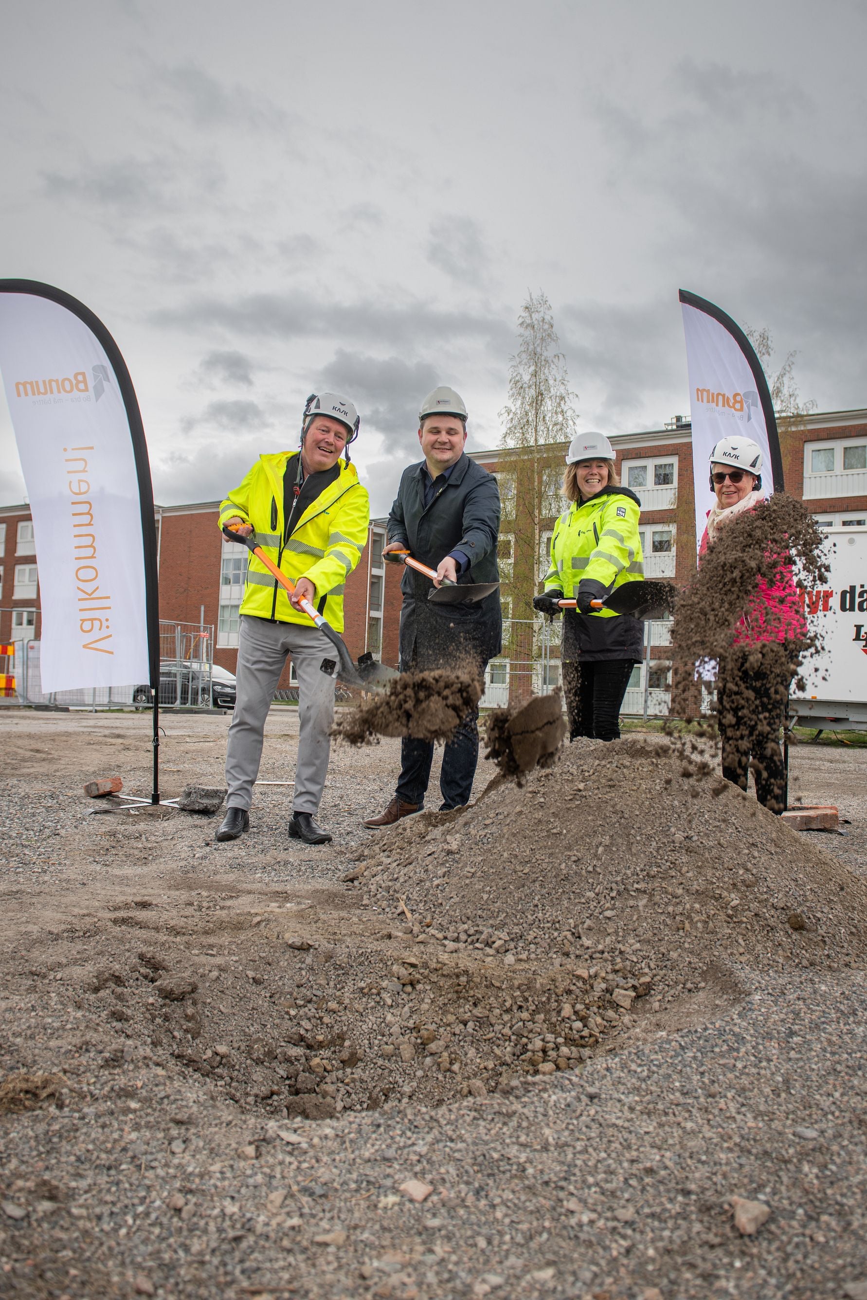 Fyra personer i reflexkläder och hjälmar deltar i en ceremoni för första spadtaget på en grusad byggarbetsplats, med bostadshus och flaggor i bakgrunden