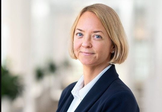 Kvinna med blont kort hår klädd i en mörk kavaj och vit skjorta, fotograferad i en ljus inomhusmiljö