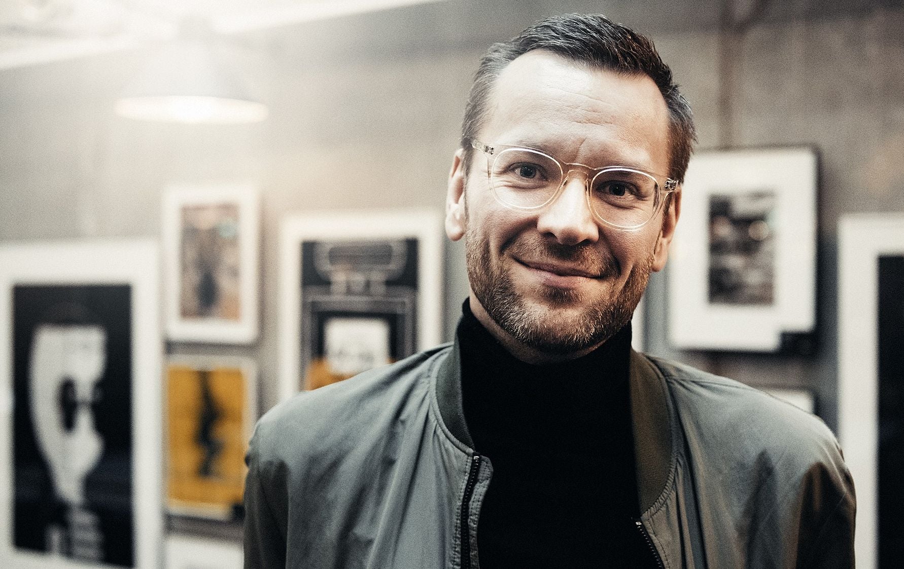 Person i en grå jacka och svart polotröja står framför en vägg med inramade bilder i bakgrunden