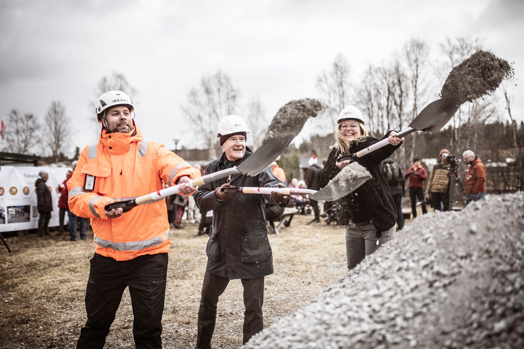 Tre personer med hjälmar och spadar skottar grus vid en markceremoni utomhus, omgivna av fler människor och träd i bakgrunden
