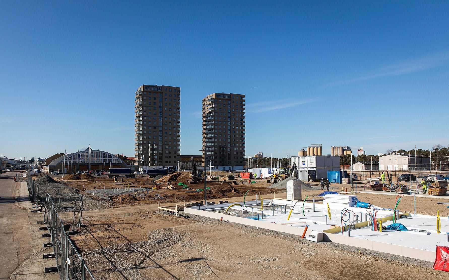 Byggarbetsplats med flerbostadshus i bakgrunden, byggmaterial och utrustning på marken under en klar blå himmel