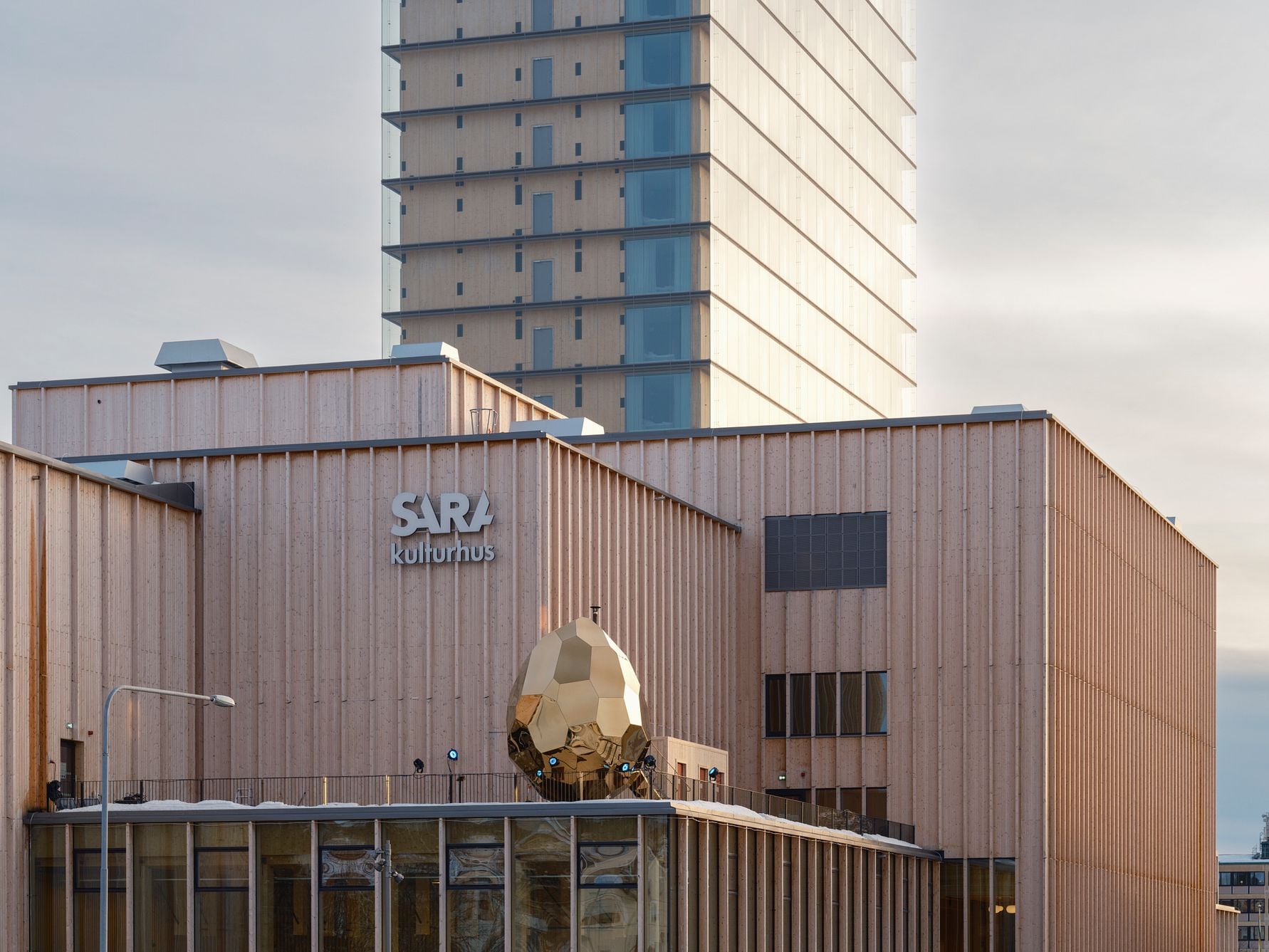 Sara kulturhus med en modern träfasad och ett högt torn i bakgrunden under en ljus himmel