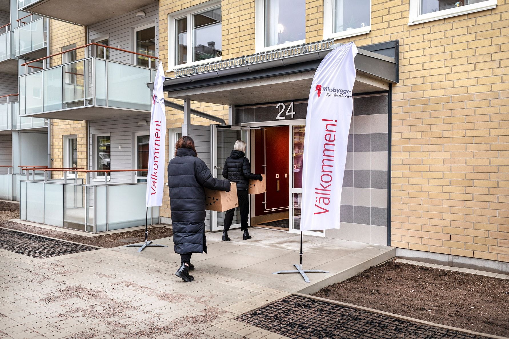 Personer går in i en modern byggnad med balkonger, där två flaggor med texten "Välkommen!" står vid entrén