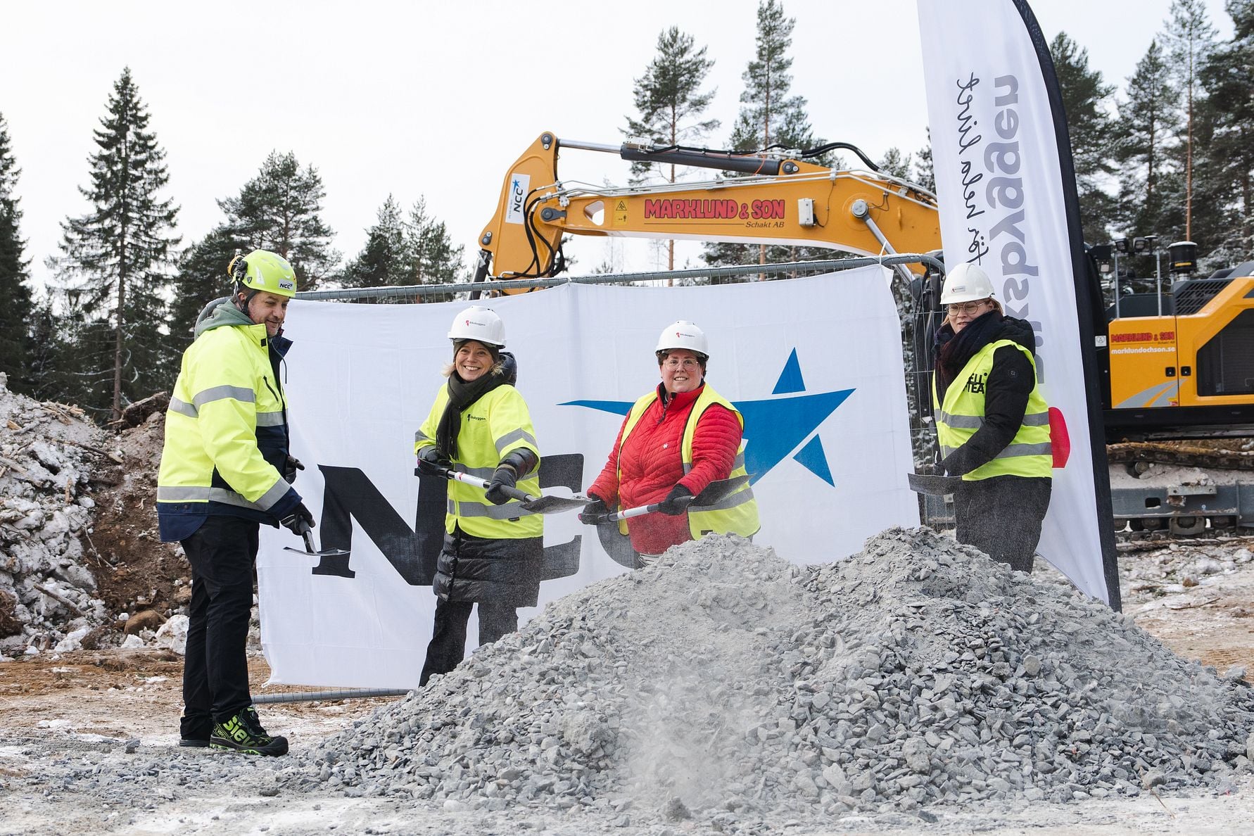Personer med skyddsutrustning vid en byggarbetsplats, framför en grushög och en grävmaskin, med en företagsbanner i bakgrunden