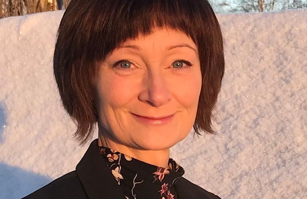 En person med mörkt hår iklädd en svart jacka och en blommig blus står utomhus med en snöig bakgrund och soligt ljus