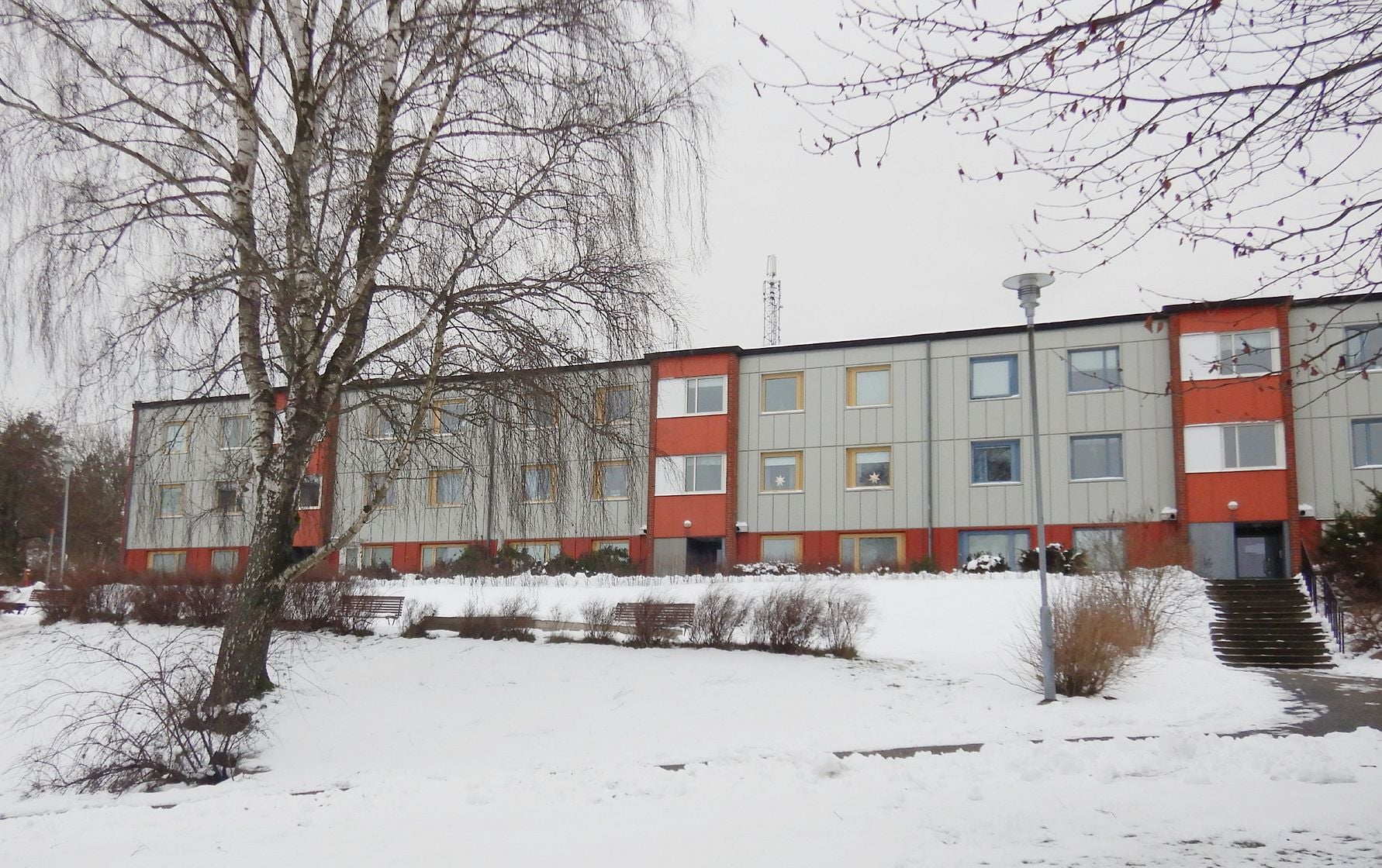 Trebånings flerfamiljshus med plåtfasad i grått och orange, omgivna av snöklädd mark och ett träd i förgrunden