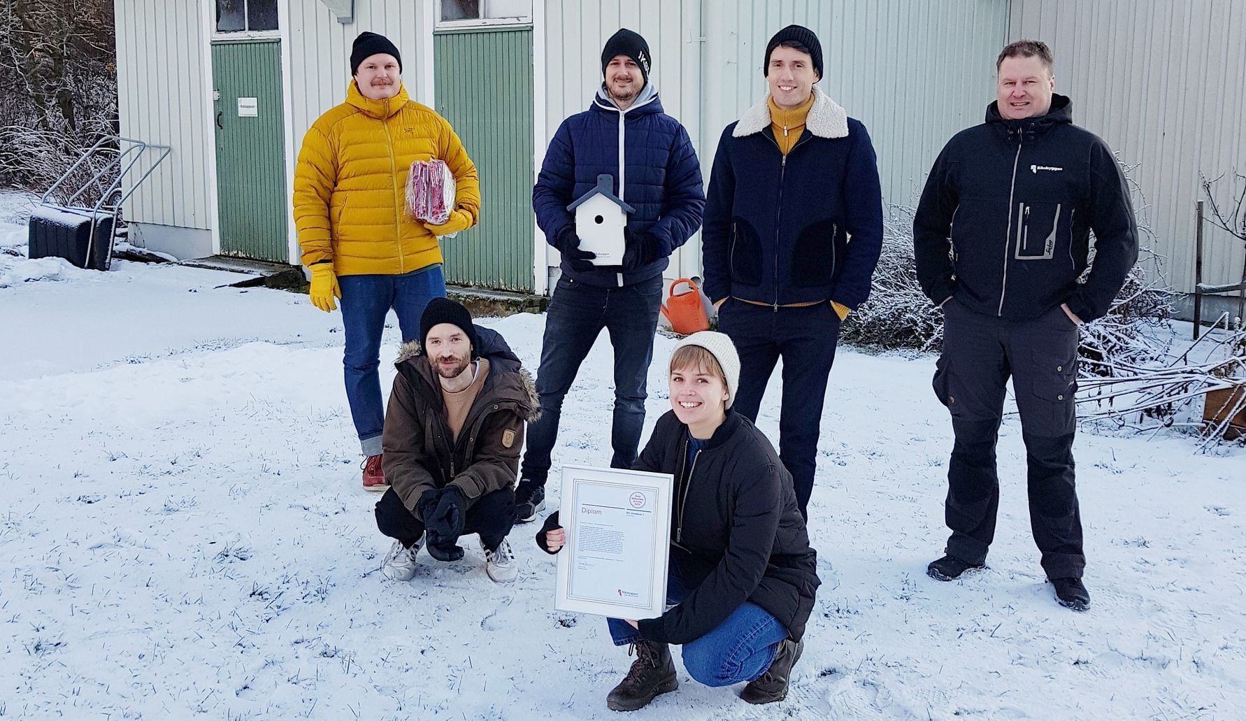 En grupp människor står utomhus på en snötäckt mark framför en byggnad, vissa håller föremål såsom ett diplom, en fågelholk och ett paket