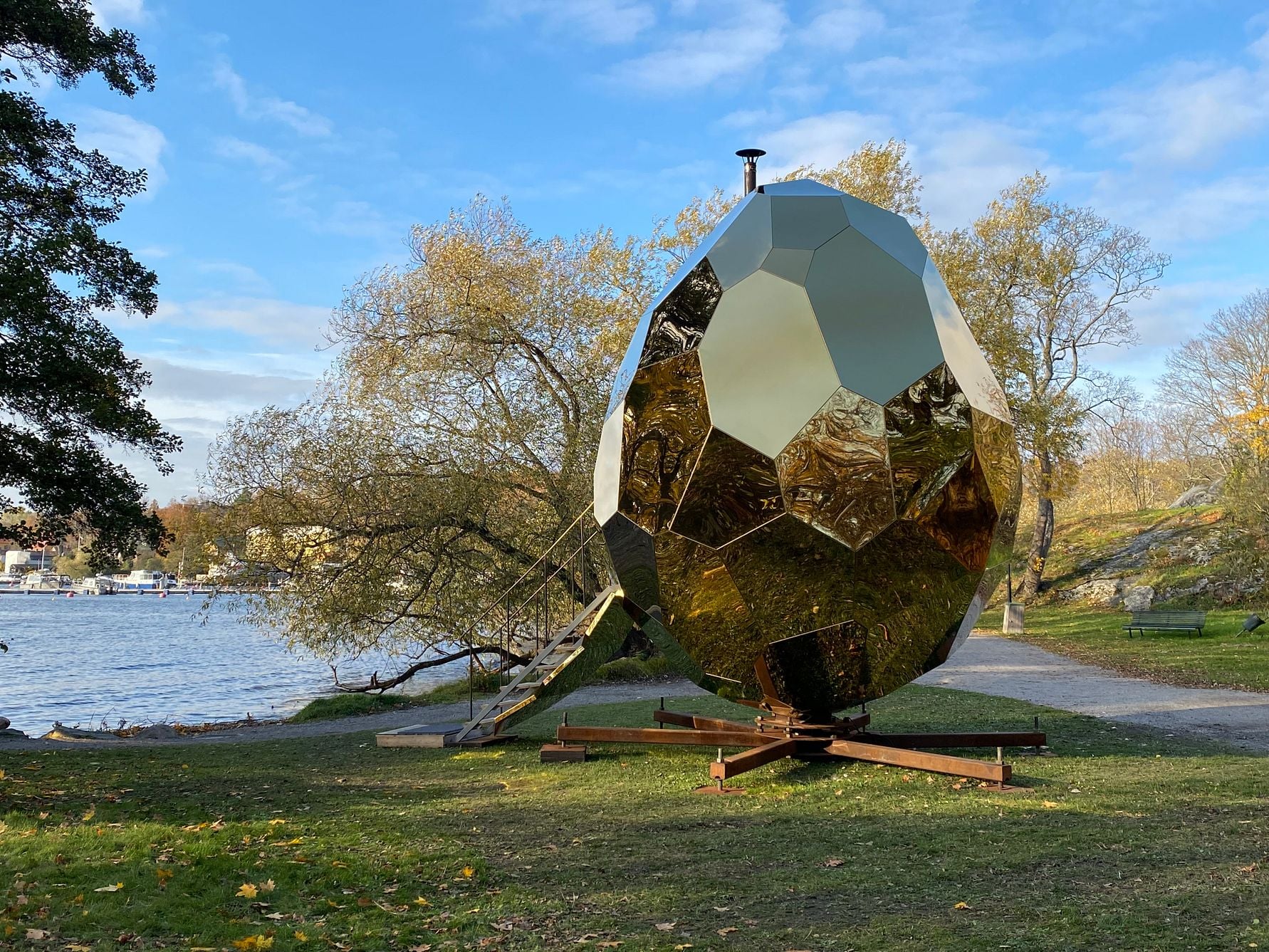 Guldfärgat polygonformat konstverk utomhus vid en park bredvid en sjö, omgiven av höstträd och blå himmel